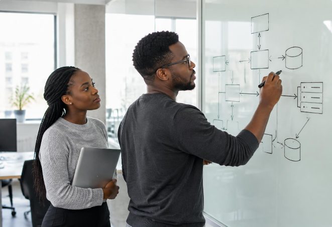 Two colleagues discussing a system diagram drawn on a whiteboard in an office